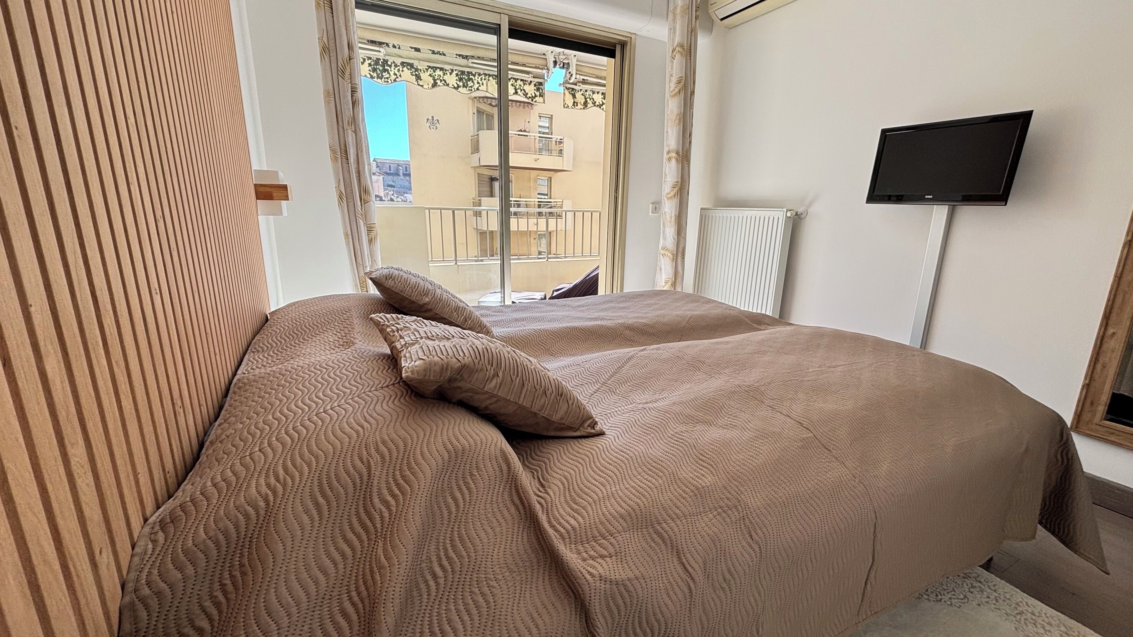A hotel room with a bed, a television on the wall, and a window with a view of buildings.