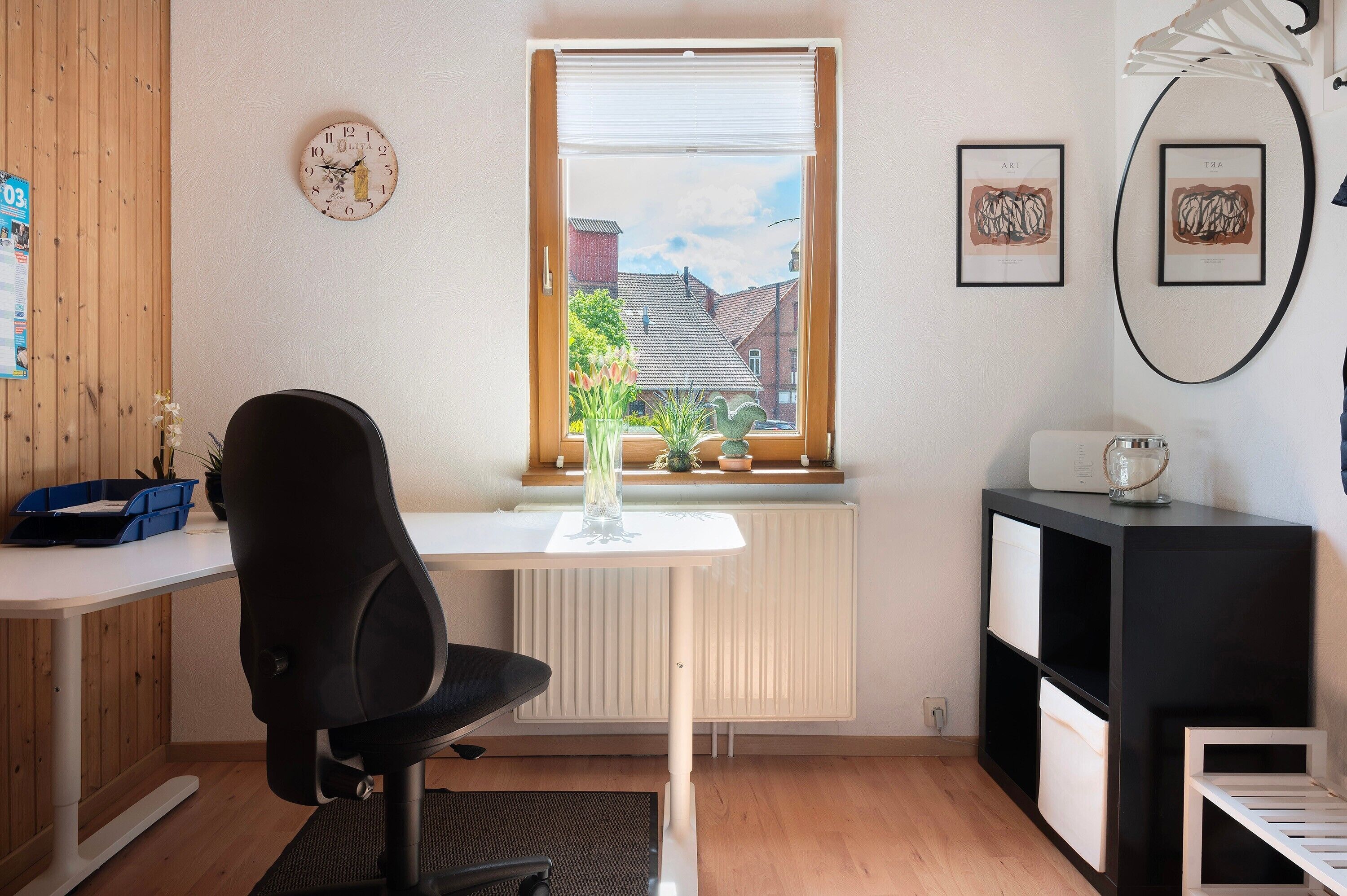 A room with a desk, chair, radiator, window with blinds, framed artwork, and a small shelf with a decorative item.