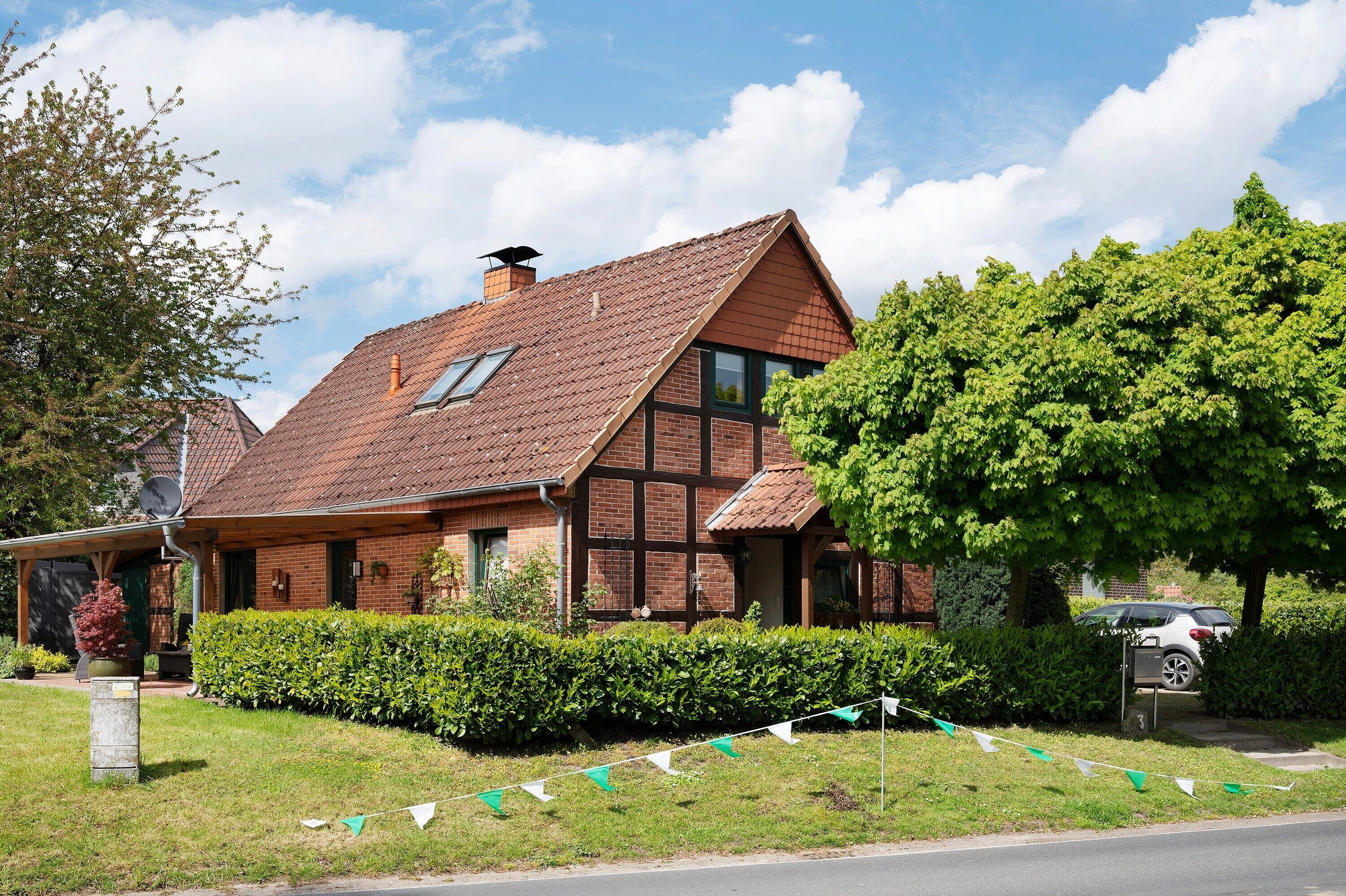 Ein traditionelles Backsteinhaus mit geneigtem Dach, einem sichtbaren Schornstein und einem gepflegten Garten mit Hecke und Baum.