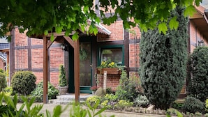Un edificio de ladrillo con una pérgola de madera, un letrero de bienvenida y una variedad de plantas y arbustos en el jardín.