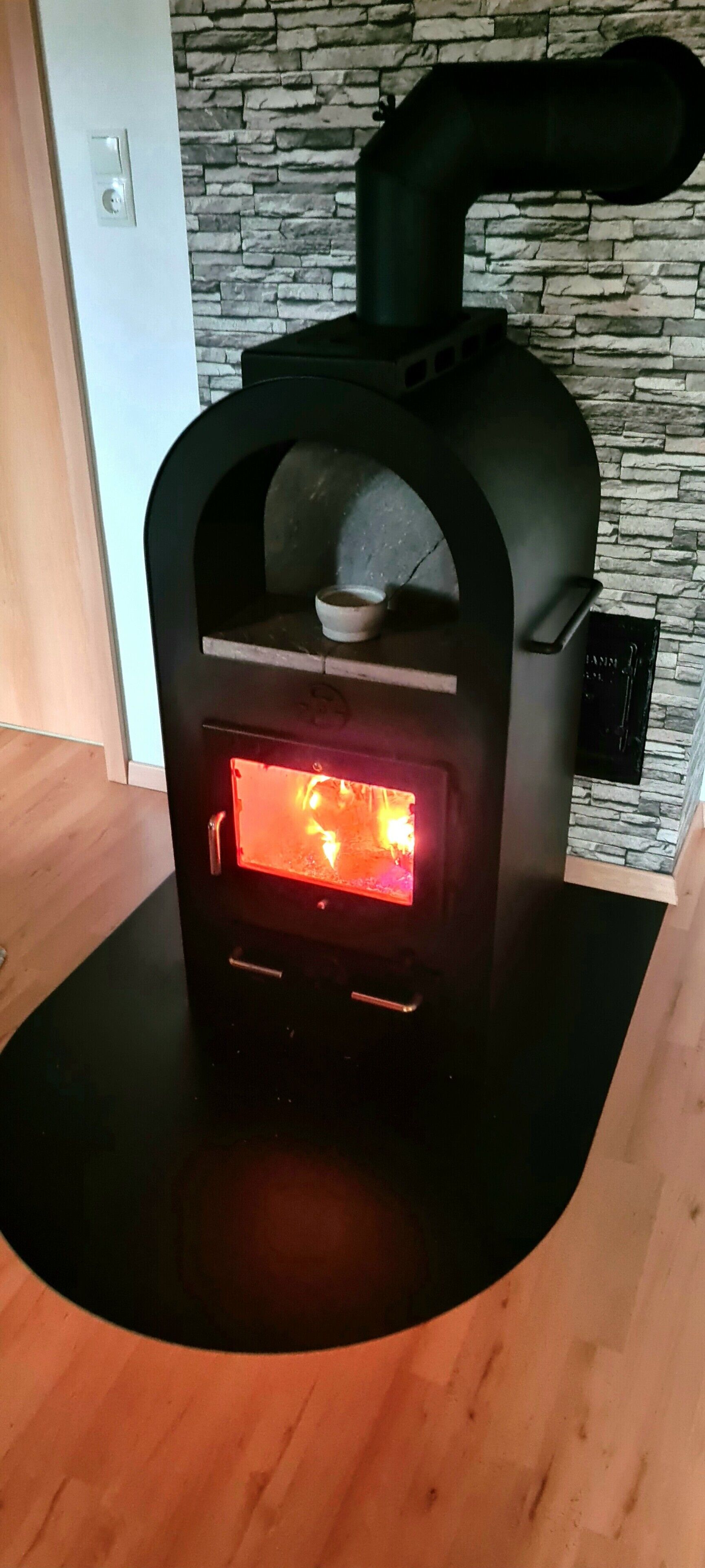 Een houtkachel met zichtbaar vuur, geplaatst op een zwarte mat, voor een achtergrond van een stenen muur.