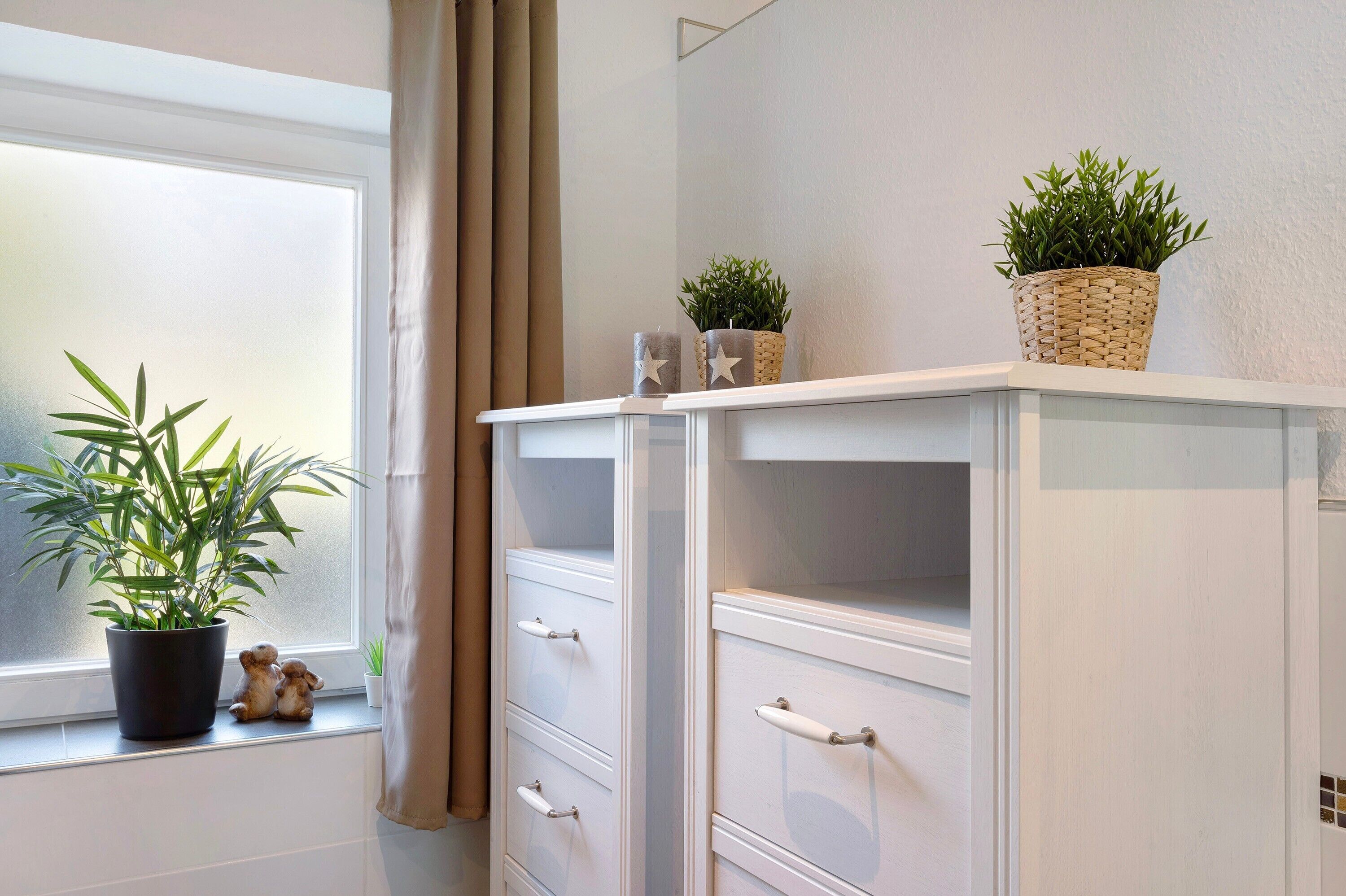 Una habitación con una ventana, un armario blanco con cajones y dos plantas en macetas sobre el alféizar.