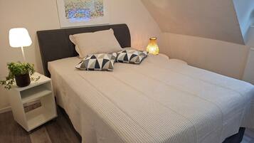 A bedroom with a bed, pillows, a nightstand with a lamp, a framed picture on the wall, and a potted plant.
