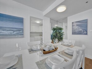 A modern dining room with a white table set for six, a kitchen in the background, and a large window with a view of the ocean. - Spacious Ocean View Condo in Beachfront Hotel Resort Amenities 1608 (Miami Beach)