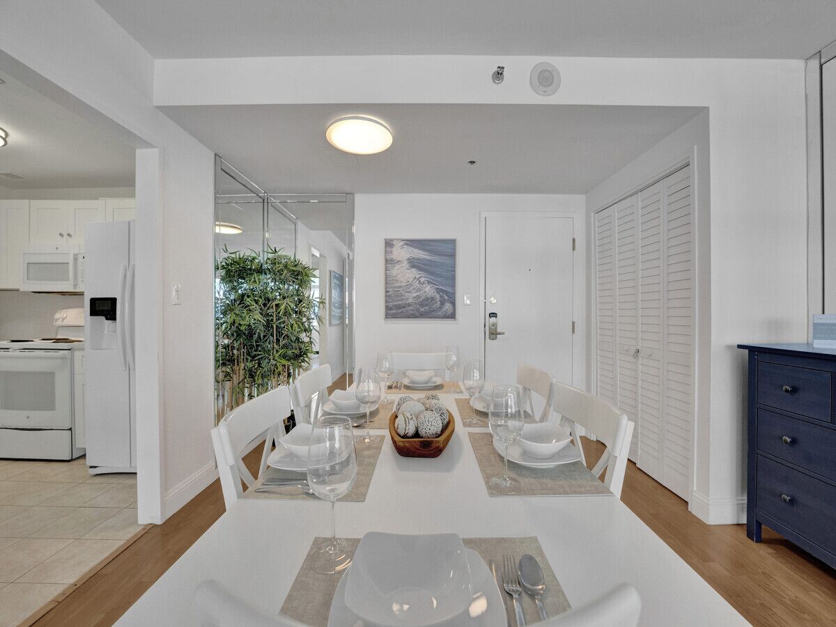 A modern dining room with a white table set for six, a kitchen in the background, and a view of the outdoors.
