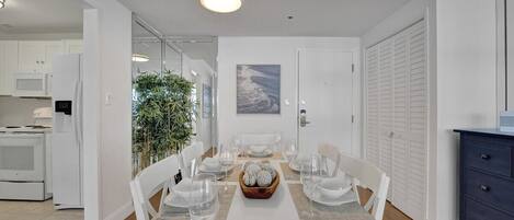 A modern dining room with a white table set for six, a kitchen in the background, and a view of the outdoors.