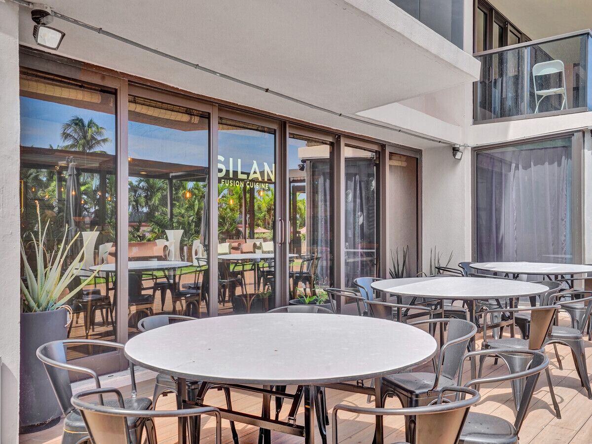 Un espace de restauration en plein air avec des tables et des chaises rondes, de grandes fenêtres et une vue sur des palmiers.