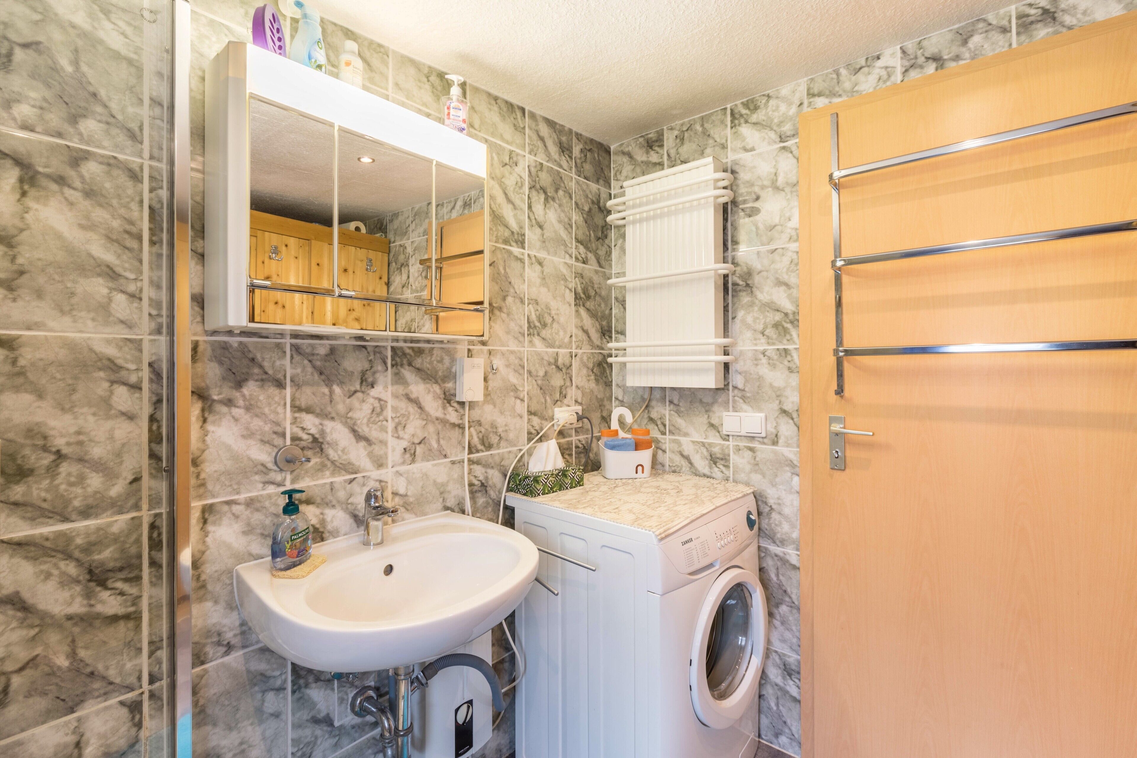 A bathroom with a marble wall, a sink, a mirror, a washing machine, and a wooden door.