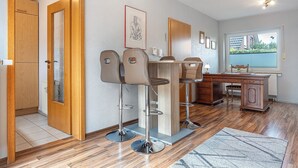 A room with a wooden door, a desk with a computer, two bar stools, and a window with blinds. - Apartment 'Akazie' with Private Terrace and Wi-Fi (Löhne)