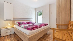 A bedroom with a wooden bed, a nightstand with a lamp, a wooden chair, and a window with blinds. - Apartment 'Akazie' with Private Terrace and Wi-Fi (Löhne)