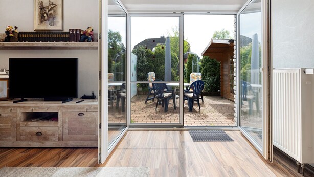 A modern living room with a television, wooden cabinet, and a glass door leading to an outdoor patio with a table and chairs. - Apartment 'Akazie' with Private Terrace and Wi-Fi (Löhne)