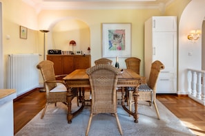 Un coin repas avec une table en bois et des chaises en osier, un radiateur, un meuble blanc et une œuvre d’art encadrée accrochée au mur.