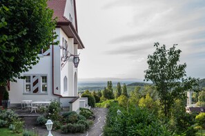 Un bâtiment blanc avec un toit rouge, un jardin avec un chemin pavé et une vue sur des collines lointaines.
