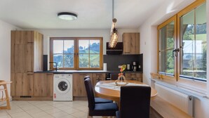 Cocina con armarios de madera, lavadora, mesa de comedor con sillas y un amplio ventanal con vistas a la vegetación.