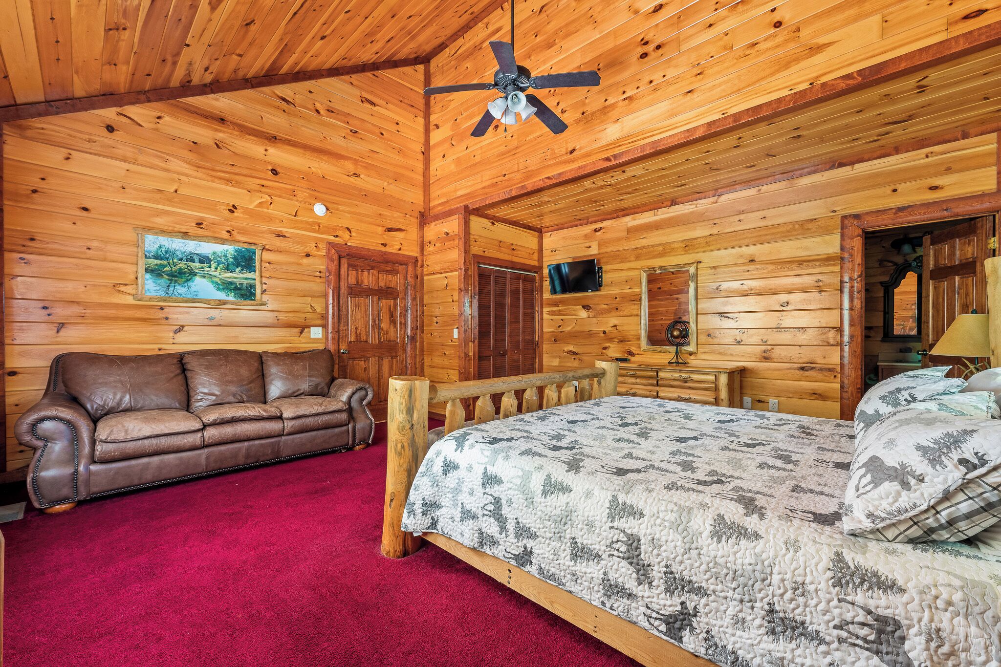 Une chambre aménagée dans un chalet en rondins de bois, avec un plafond en bois, un lit recouvert d’une courtepointe aux motifs forestiers, un canapé en cuir marron et un téléviseur fixé au mur.