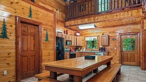 A wooden cabin interior with a large wooden table and benches, a kitchen with wooden cabinets, and a view of greenery outside.