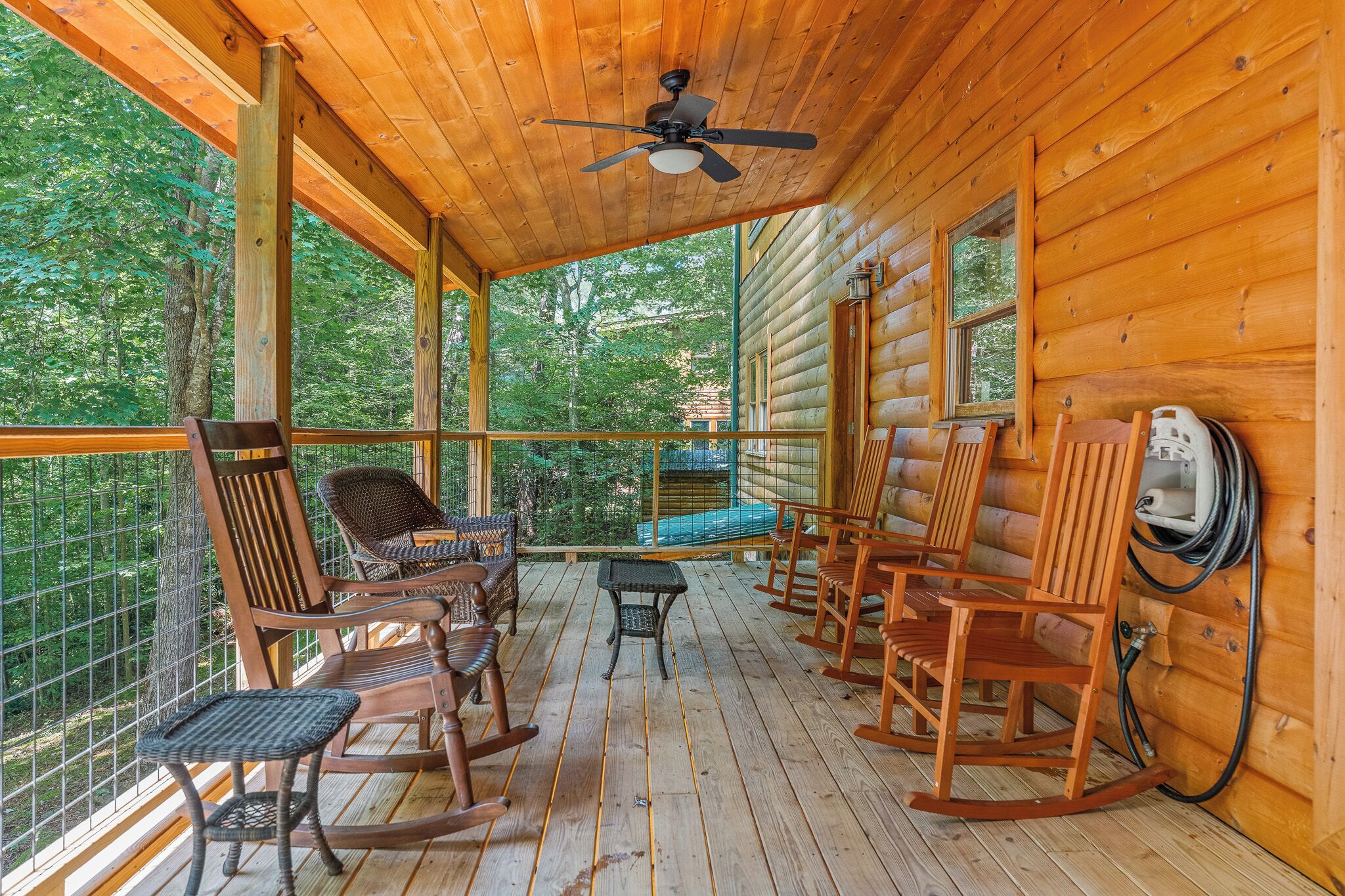 A wooden cabin with a porch featuring rocking chairs, a table, and a ceiling fan.