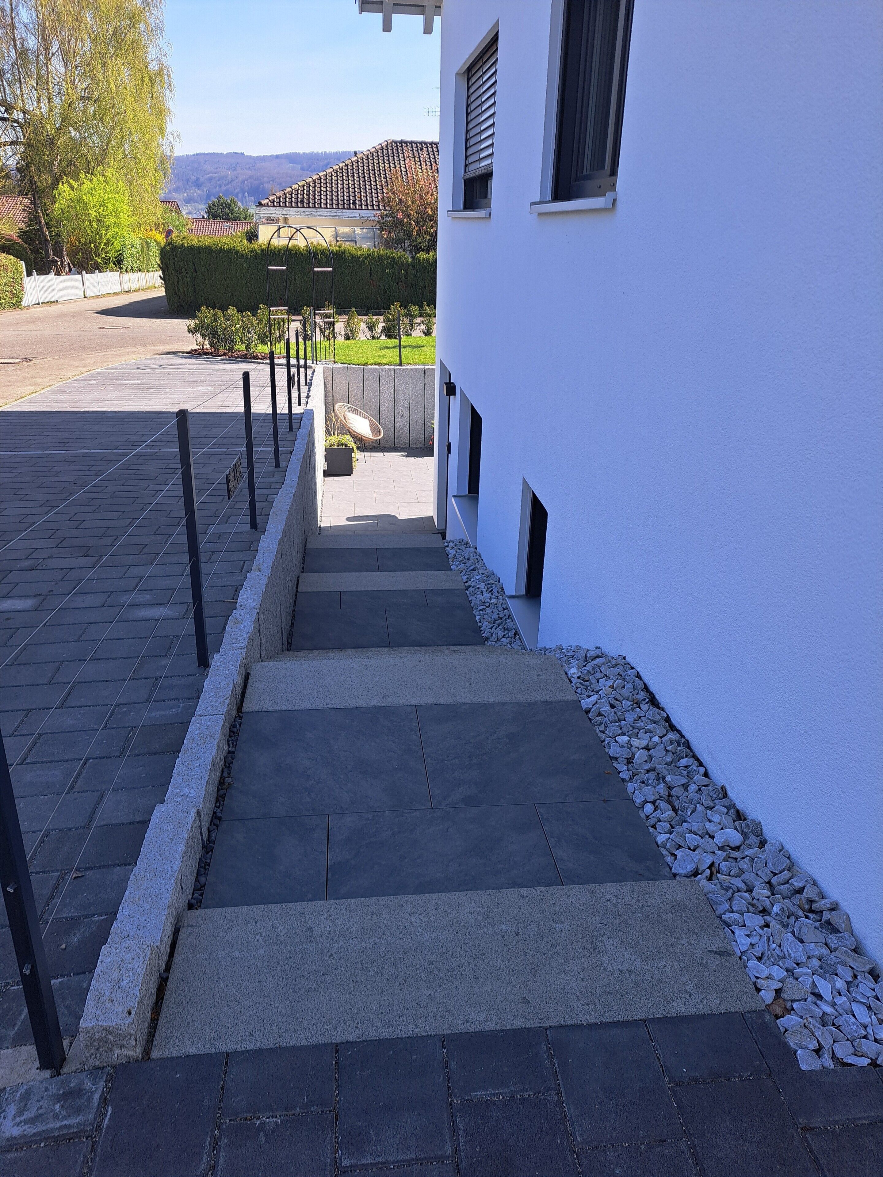 Ein Weg führt zu einem Gebäude mit weißer Fassade, einer gepflasterten Fläche und einer Aussicht auf eine grüne Landschaft im Hintergrund.