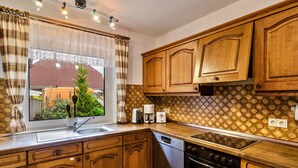 Eine Küche mit Holzausstattung, einem Geschirrspüler, einem Backofen, einem Fenster mit Vorhängen und Blick auf ein Haus.