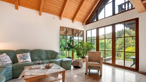 Een ruime woonkamer met een groene bank, een houten salontafel en uitzicht op een bos door grote ramen.