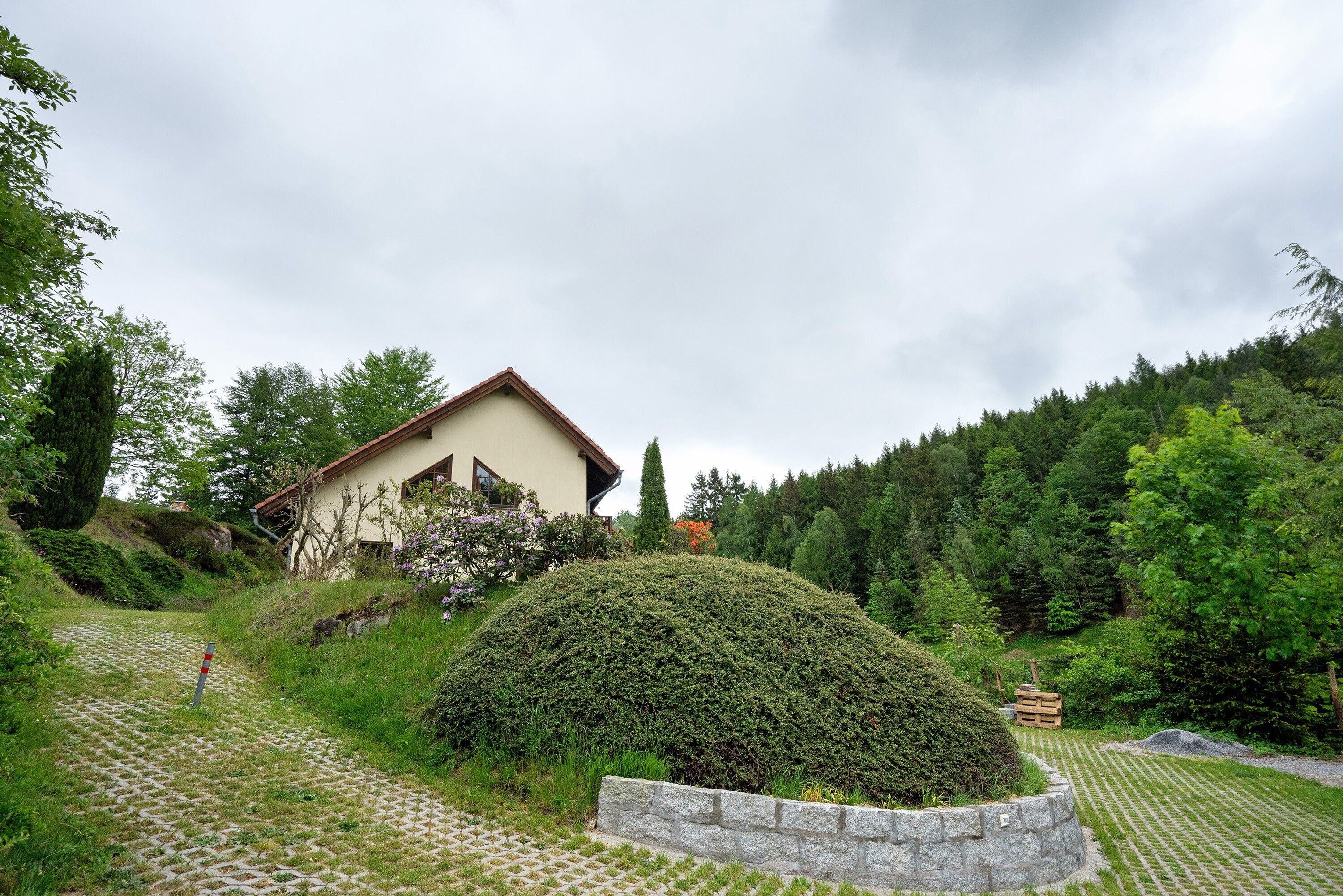A house with a sloped roof, surrounded by a well-maintained garden and a forested hillside.
