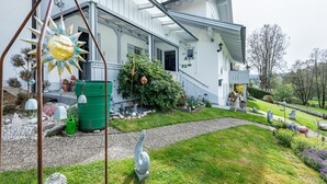 Een huis met een veranda, een groene afvalcontainer en een tuin met verschillende planten en beelden.