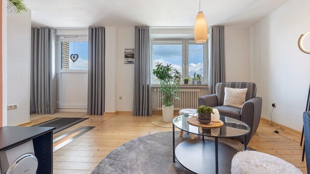A modern living room with a sofa, a glass coffee table, a white fluffy ottoman, a potted plant, and a view of the outside through large windows. - Apartment 'Weserberglandblumen' with Mountain View, Private Garden and Wi-Fi (Bad Pyrmont)