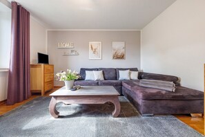 Un salon comprenant un canapé d’angle, une table basse, un téléviseur posé sur un meuble en bois et une grande fenêtre avec des rideaux.