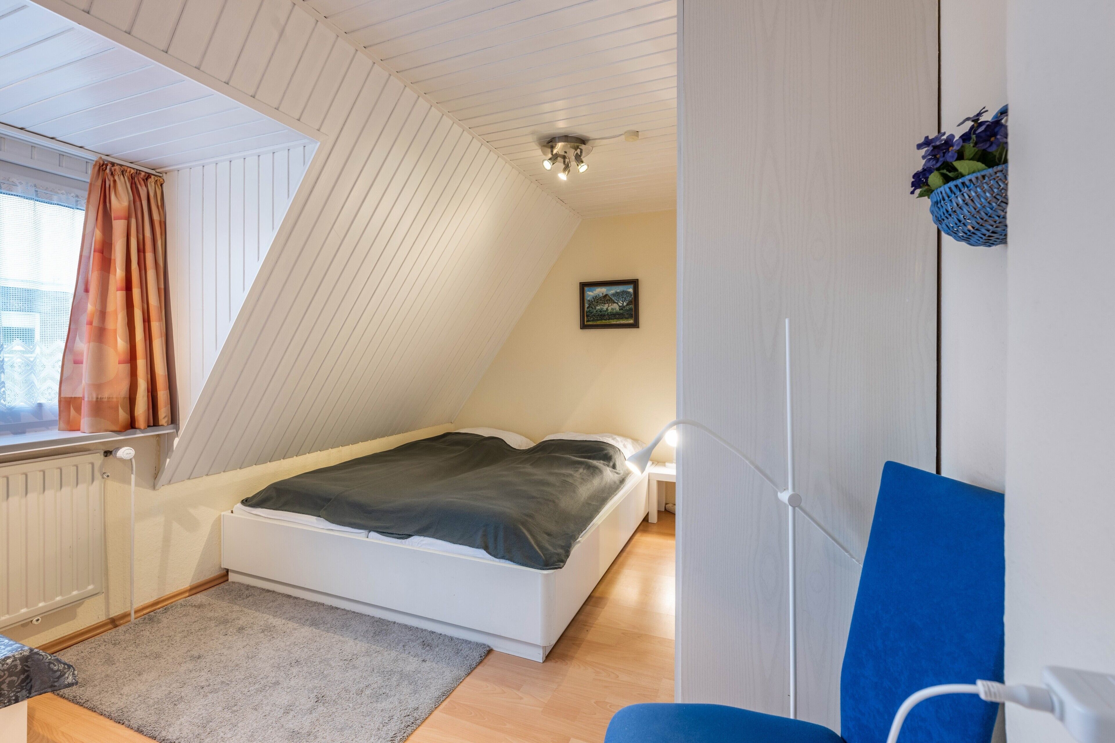 Un dormitorio con techo inclinado, una cama, una mesita de noche, una silla y una cesta con flores fijada a la pared.
