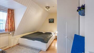 Un dormitorio con techo inclinado, una cama, una mesita de noche, una silla y una cesta con flores fijada a la pared.