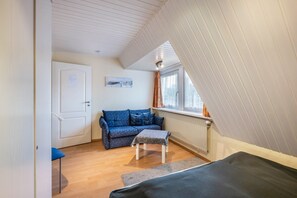 Une pièce avec un plancher en bois, un canapé bleu, une petite table, un lit, une porte blanche et une fenêtre avec des rideaux.