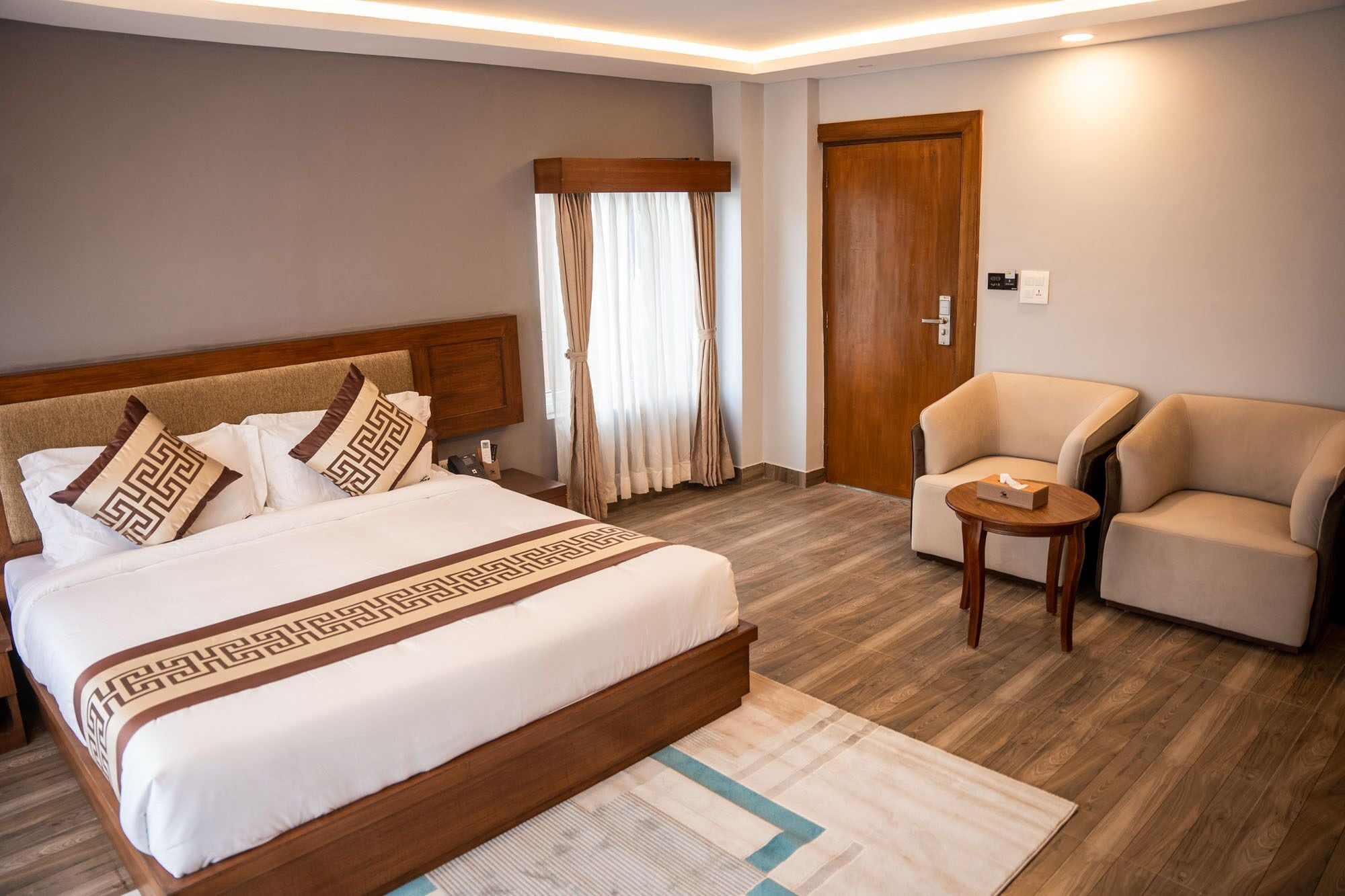 Habitación de hotel con una cama grande, cabecera de madera, puerta de madera, un sillón color beige y una mesita pequeña.
