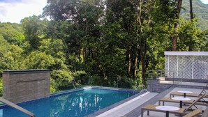 A pool area with a clear blue pool, surrounded by lush greenery and trees. There are lounge chairs and a small table near the pool. - Nagarjun Resort (Tarakeshwar)