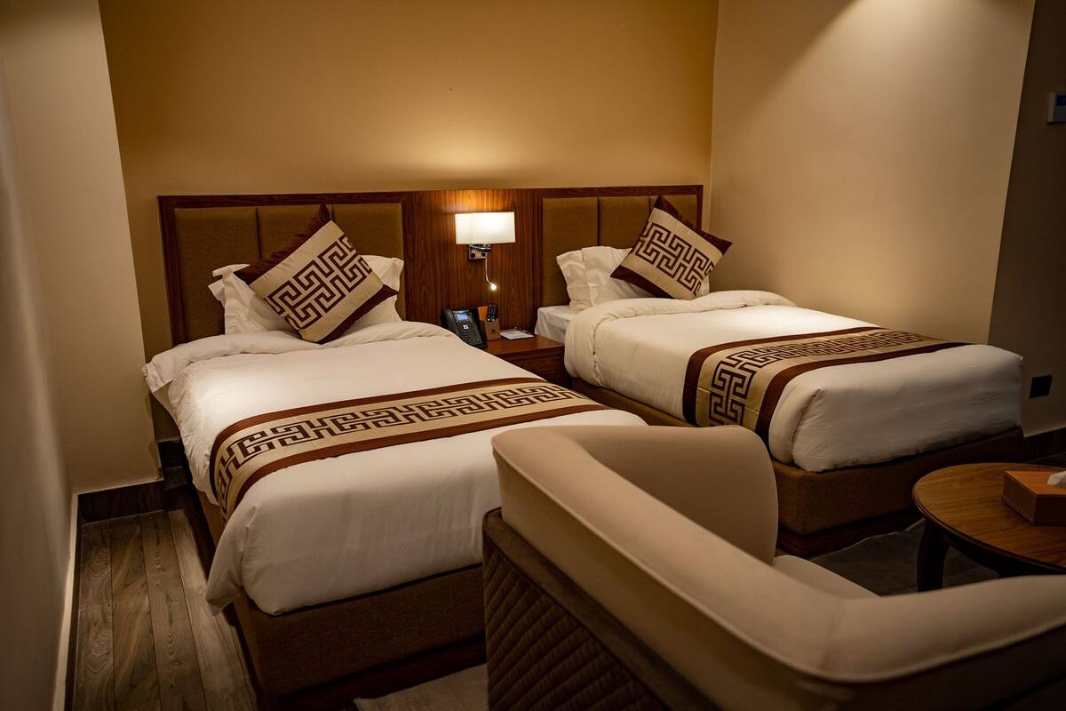 Habitación de hotel con dos camas, cada una con cabecera y almohada a juego. Hay una mesita de noche con lámpara y teléfono.