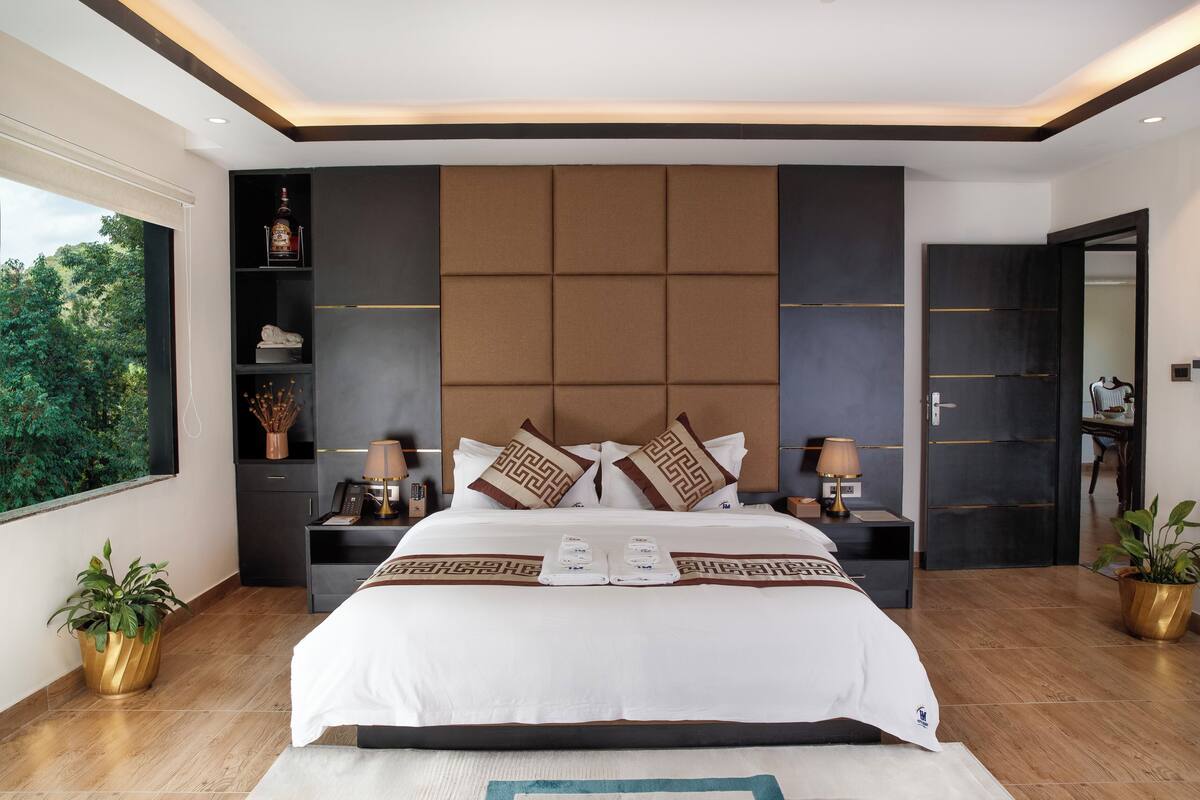 Una habitación de hotel moderna con una cama grande, cabecera integrada, mesitas de noche y una ventana con vistas a la vegetación, además de plantas en macetas.