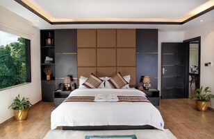 A modern hotel room with a large bed, a built-in headboard, bedside tables, a nightstand, a window with a view of greenery, and potted plants.