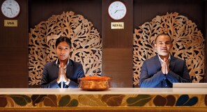 Due persone sedute allo sportello della reception con uno sfondo decorativo e orologi che mostrano i fusi orari degli Stati Uniti e del Nepal.