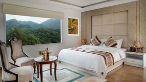 A hotel room with a large bed, a sitting area with a table, and a view of a forested mountain range. - Nagarjun Resort (Tarakeshwar)