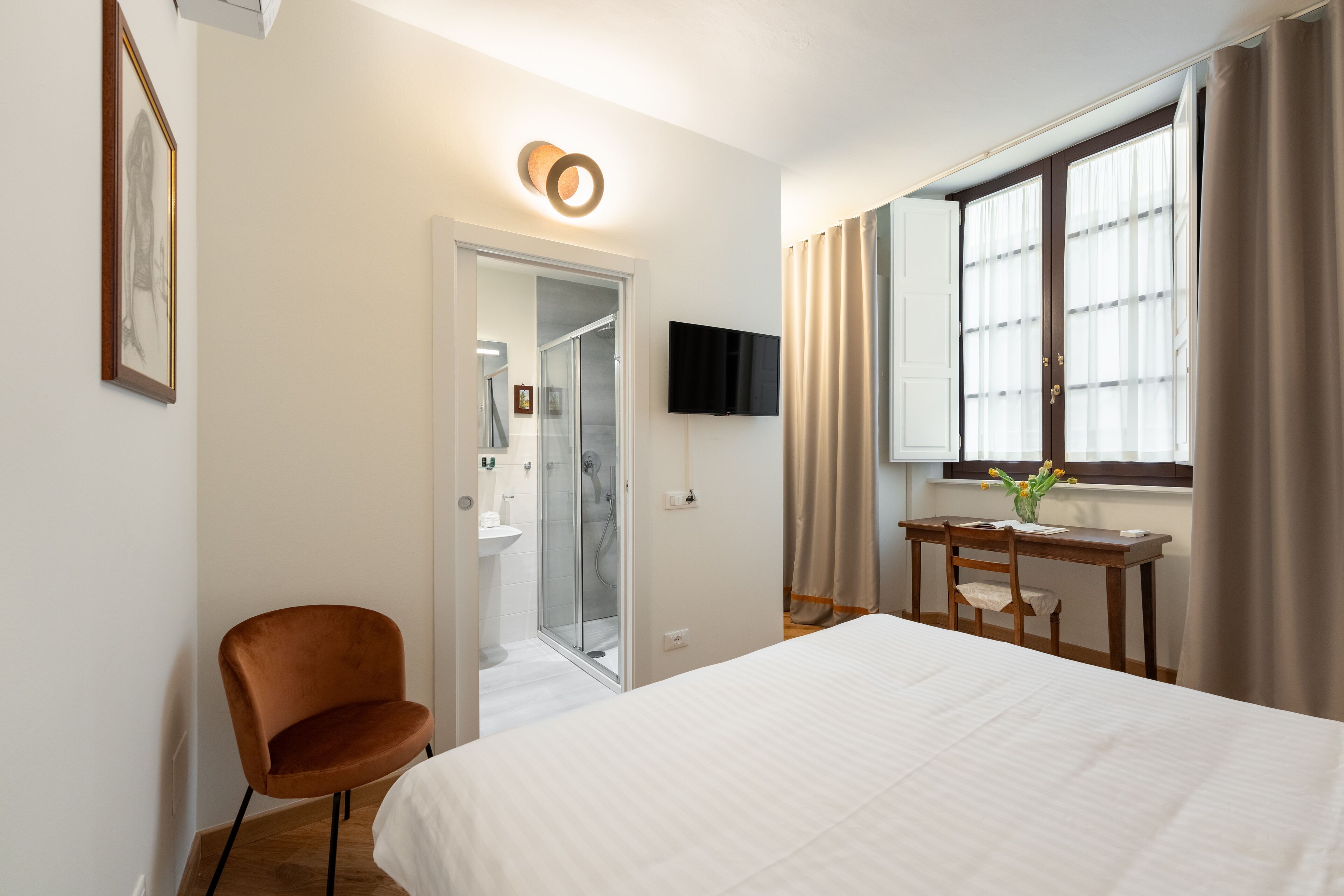 Une chambre d’hôtel comprenant un lit, un bureau avec une chaise, une télévision, une douche et une fenêtre avec des rideaux.