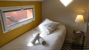 En säng med plats för en person, täckt med vita sängkläder, ett fönster med utsikt över en röd byggnad, en sänglampa och ett mönstrat golv.