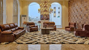 A spacious lobby with leather sofas, a patterned rug, a chandelier, and a view of the city through large windows. - Hotel S Crown Somnath  (Veraval)