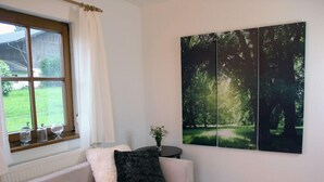 Una sala de estar moderna con un sofá blanco, una ventana con cortinas, un jarrón con flores y una gran obra de arte con temática forestal en la pared.