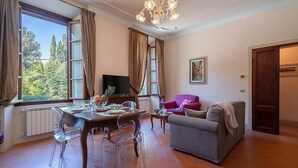 A spacious living room with a dining area, large windows, a chandelier, and a cozy seating arrangement.