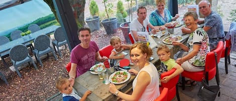 A group of people dining at an outdoor table with chairs, a wooden table, and a view of greenery in the background.