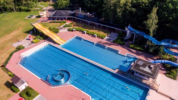 Vue aérienne d’une grande piscine avec un toboggan, entourée d’un espace aménagé comprenant des allées et un bâtiment.