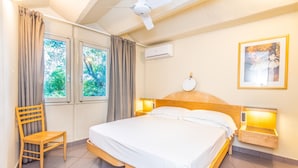 Hotelkamer met een bed, een stoel, een plafondventilator, een raam met gordijnen, een airconditioning en een nachtkastje met lamp, plus een ingelijste foto aan de muur.
