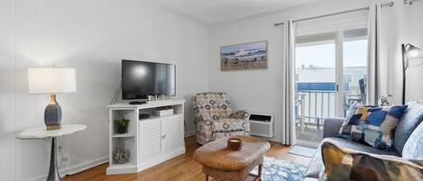 A living room with a flat-screen TV, a white entertainment unit, a wicker ottoman, a patterned sofa, and a view of the outdoors through a sliding glass door.