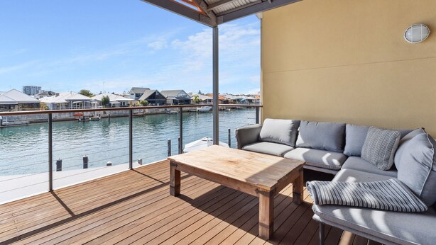 Una terraza con una mesa de madera y un sofá gris con cojines a rayas, con vista a un puerto deportivo con barcos y edificios.