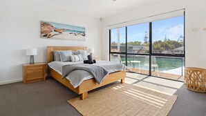 Un dormitorio moderno con una cama grande, una mesita de noche, una lámpara, un cuadro y vista al agua a través de amplias ventanas.