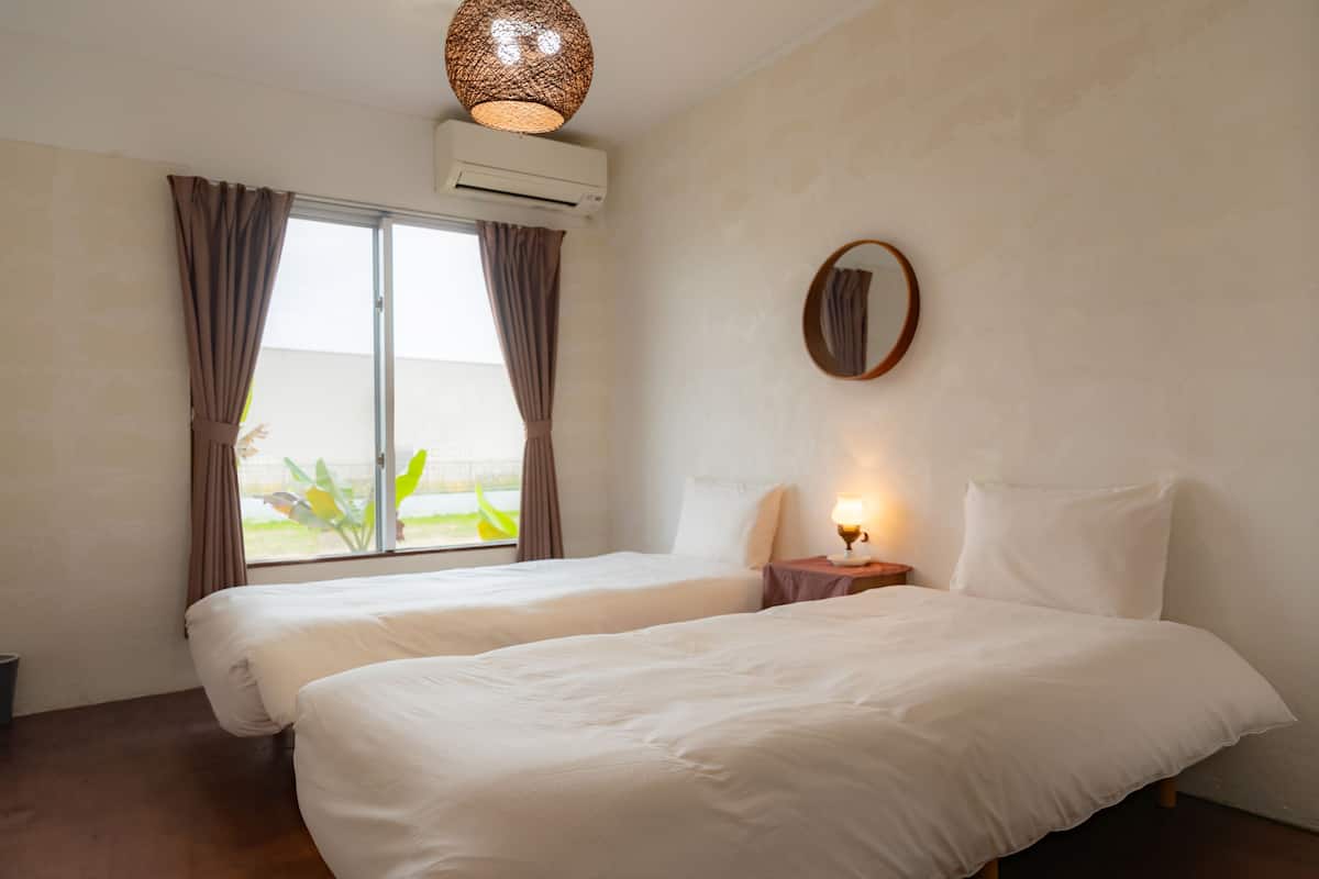 A hotel room with two beds, a window with curtains, an air conditioning unit, a bedside table with a lamp, and a round mirror on the wall.